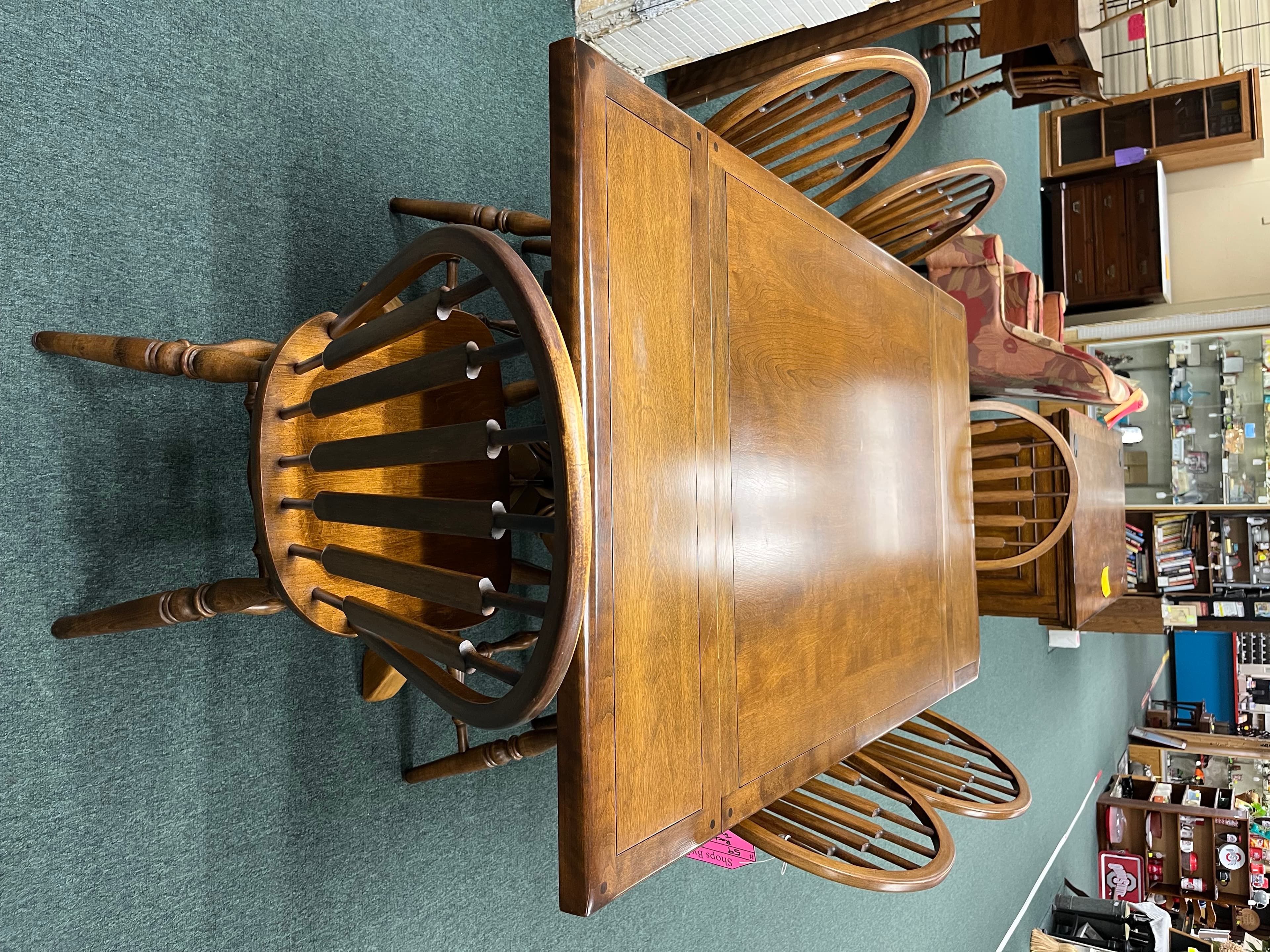 A maple dining table surrounded by Windsor-style chairs, lit by a teal-green showroom floor.