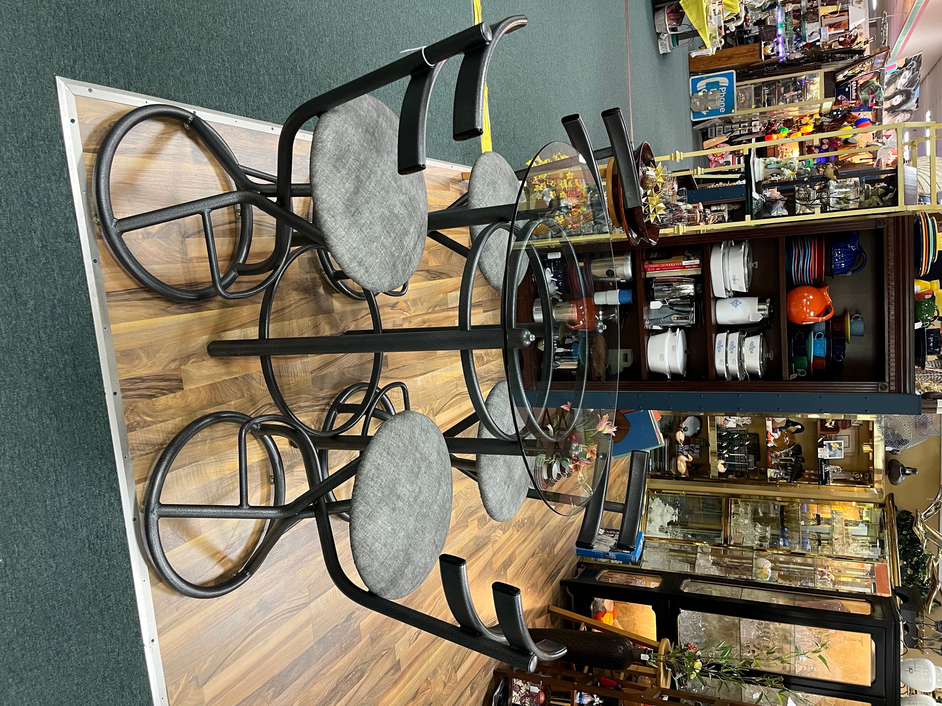 A round glass-topped bistro table with four matching iron chairs and grey cushions.